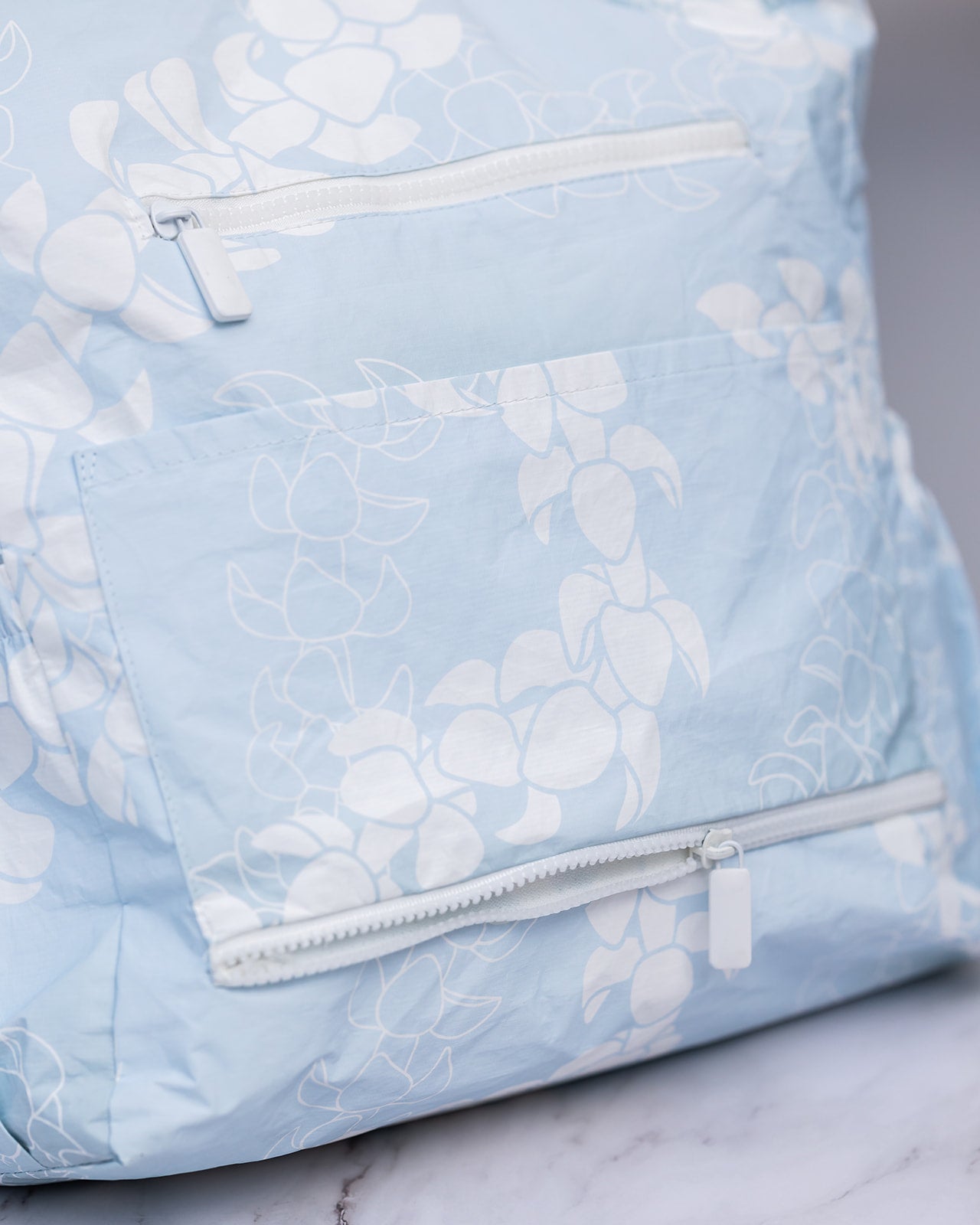Close up of Puakenikeni tyvek tote bag showing a zippered pocket and an additional zippered luggage trolley sleeve.