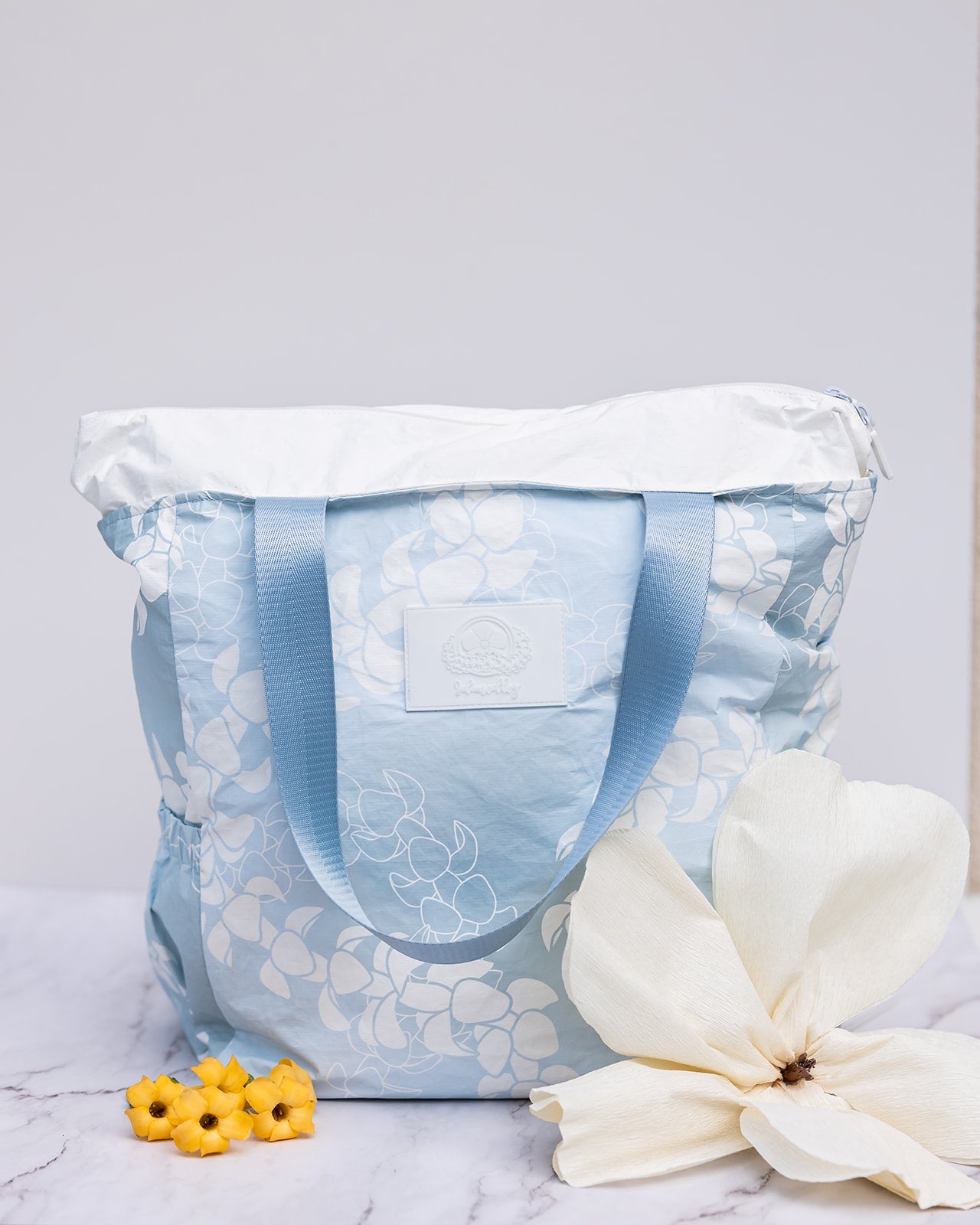 Light blue puakenikeni lei tyvek tote showing expandable zippered top and wide, matching colored nylon straps.