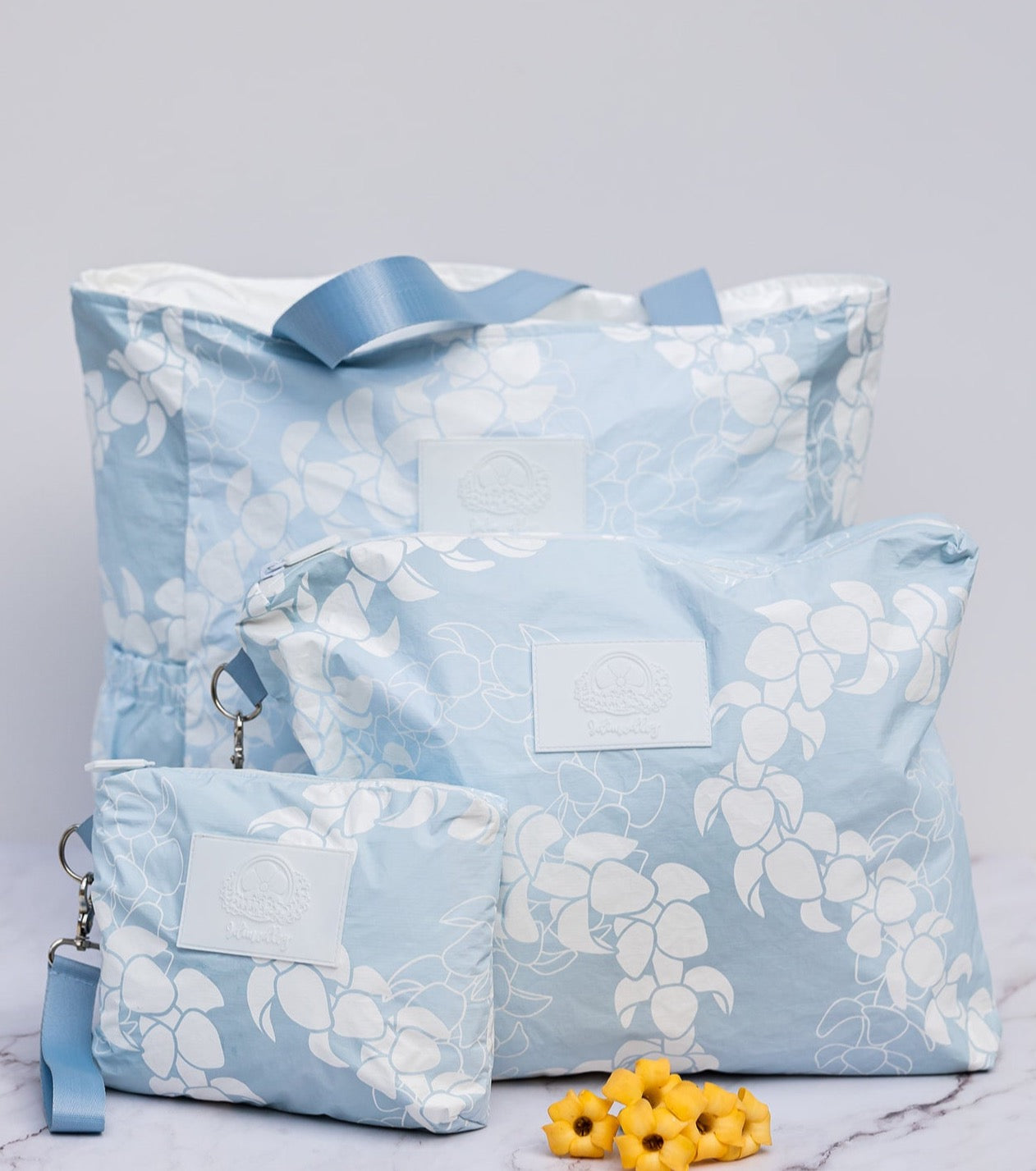 Light blue and white puakenikeni flower lei design on tyvek tote bag and matching wristlet accessory pouches of varying sizes.
