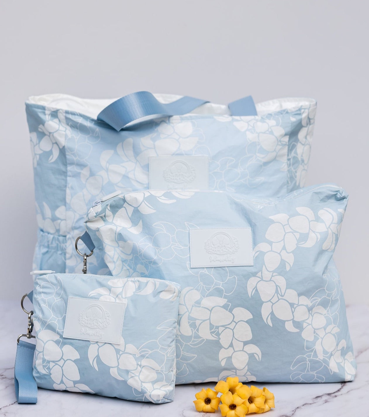 Light blue and white puakenikeni flower lei design on tyvek tote bag and matching wristlet accessory pouches of varying sizes.  