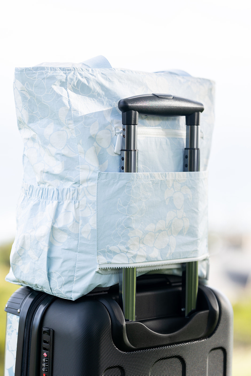 Puakenikeni lei tyvek tote sitting on luggage, held in place by luggage trolley.