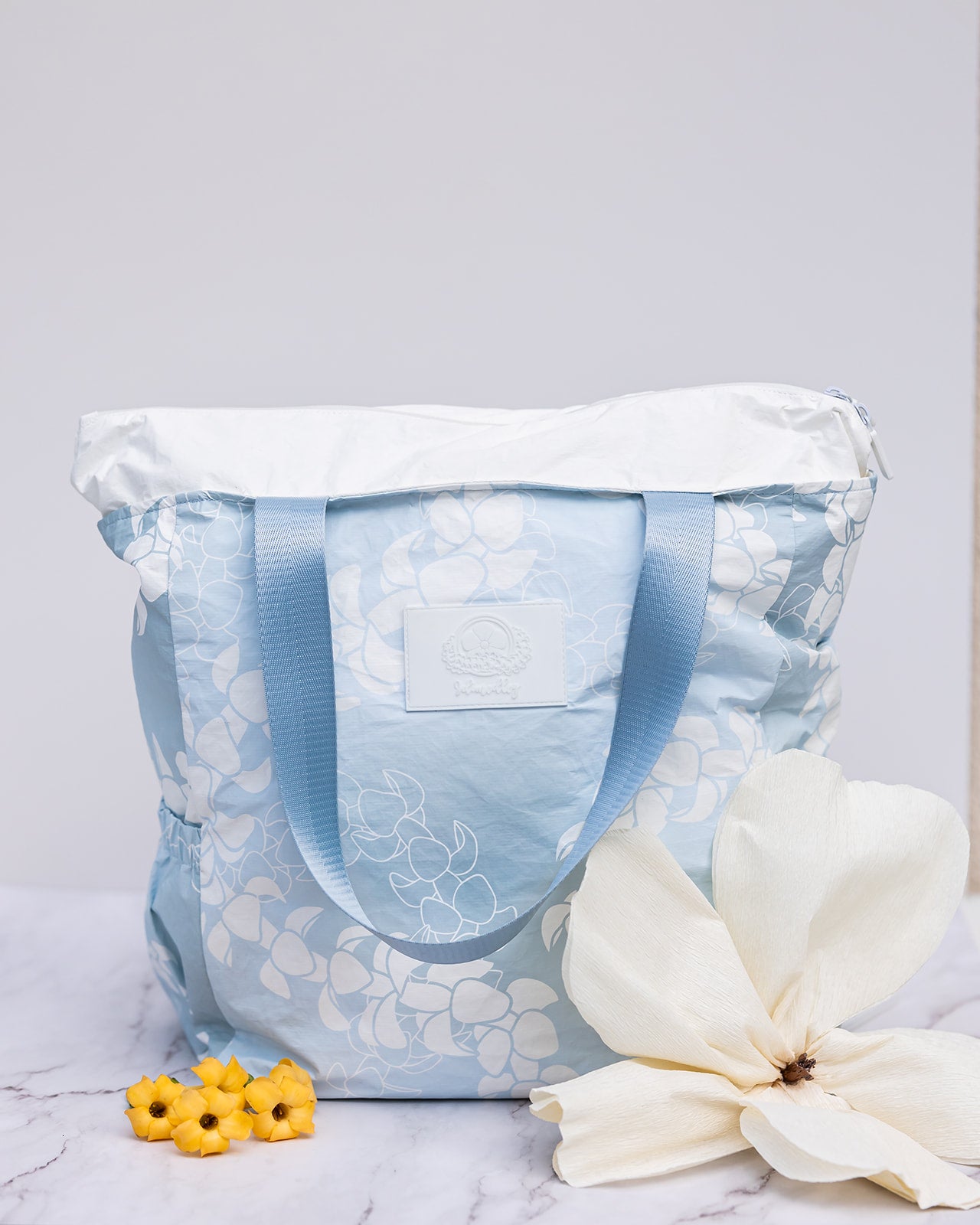 Light blue puakenikeni lei tyvek tote showing expandable zippered top and wide, matching colored nylon straps. 