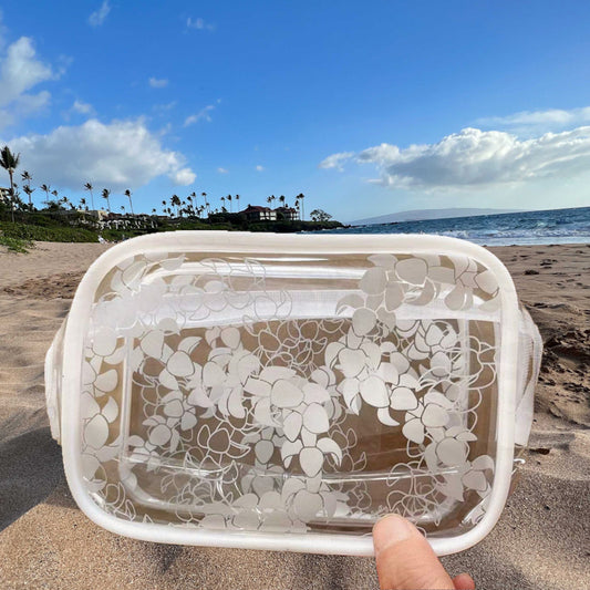 Puakenikeni_flower_lei_clear_zippered_pouch_cosmetic_bag_held_on_sandy_beach_with_blue_skies.