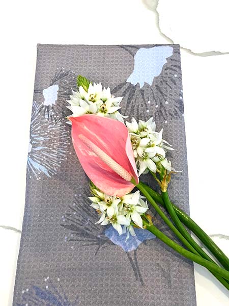 gray dishtowel with gray and periwinkle Ohia Lehua design with  pink anthurium and white flowers laid on top