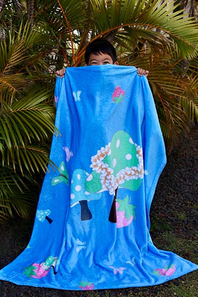 bright blue plush blanket with cute mushroom wearing plumeria lei with butterflies and strawberries design held up by smiling asian boy in front of Areca palms