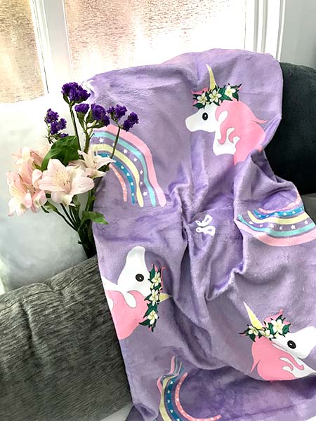 lavender plush blanket with design of unicorn wearing haku lei, butterflies and rainbows laid across gray and white pillows and pretty flowers
